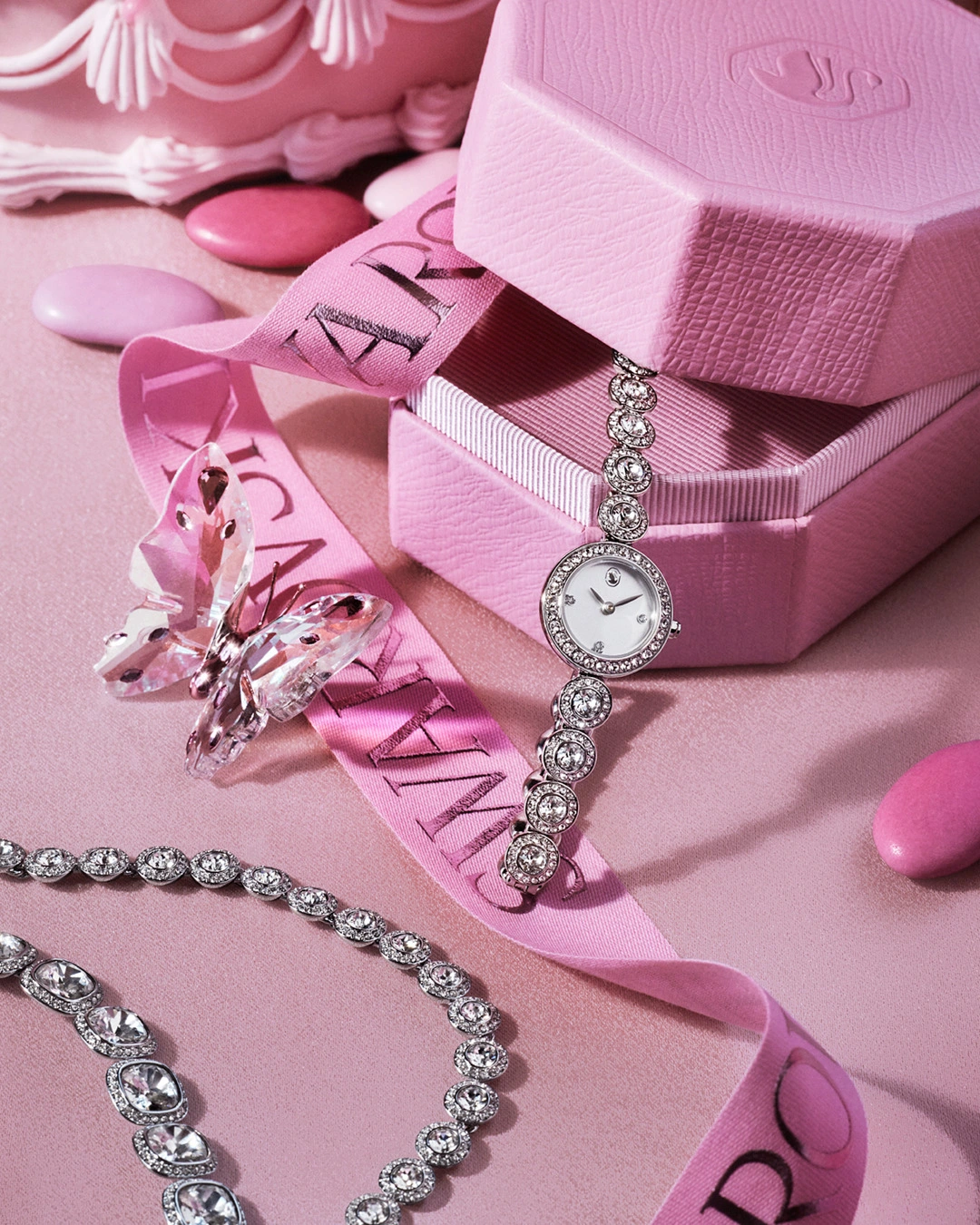Composição Swarovski com relógio e colares de cristais prateados, borboleta de cristal, caixas e fitas cor-de-rosa.